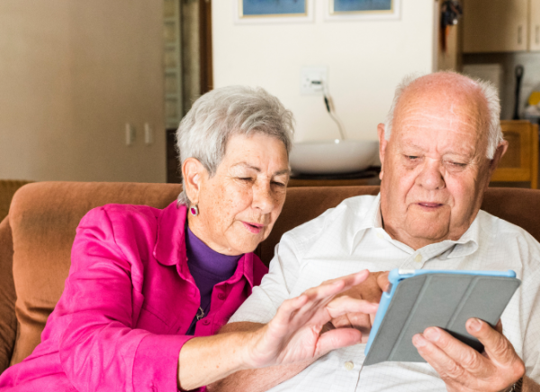 An older man and woman looking at an iPad. The woman on left is wearing a bright pink jacket. The man on the right is wearing white,