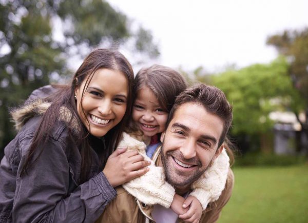 Happy family in a park. Mum and dad with a four year old daughter. A little girl is riding on hear dad's back.